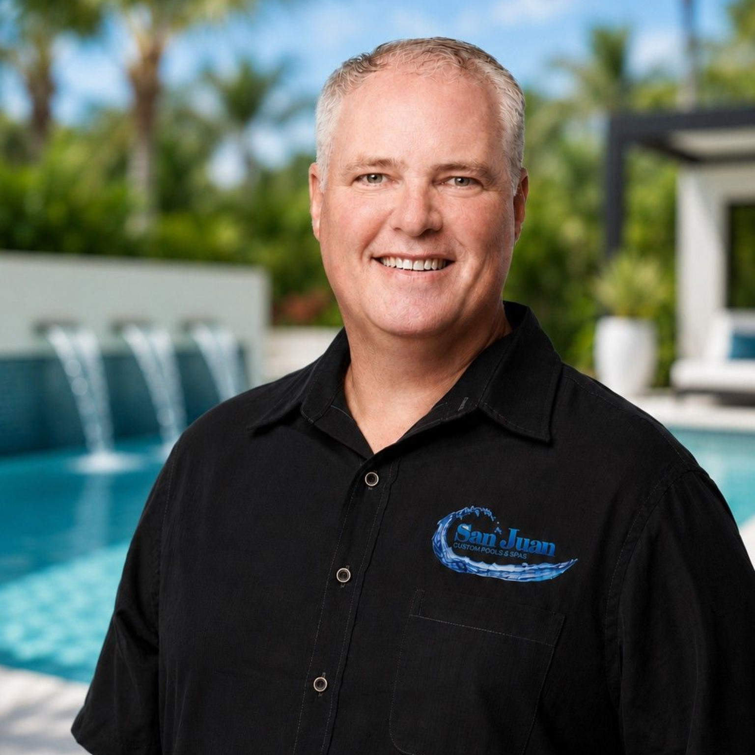 Smiling man in black shirt standing by a pool with a waterfall in the background