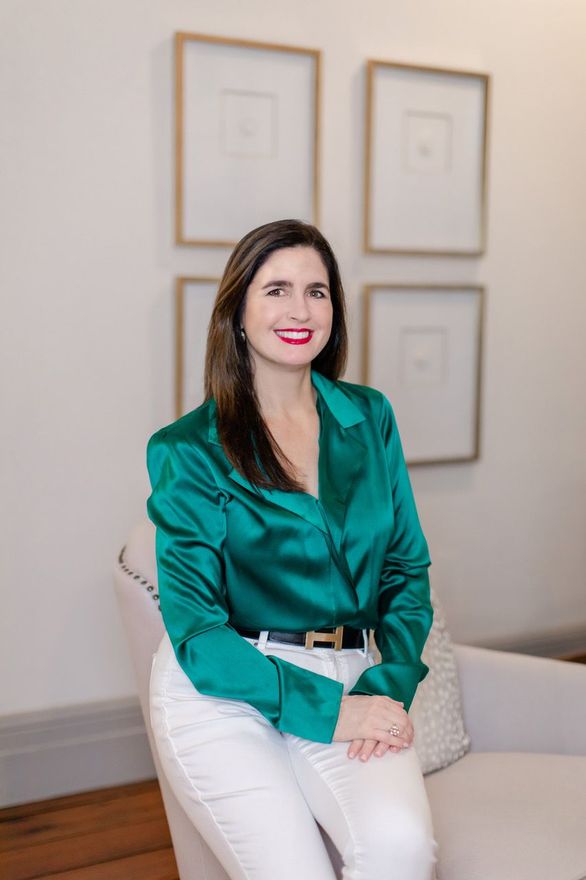 Woman seated on a chair in a teal blouse and white pants, smiling in a minimalist room.