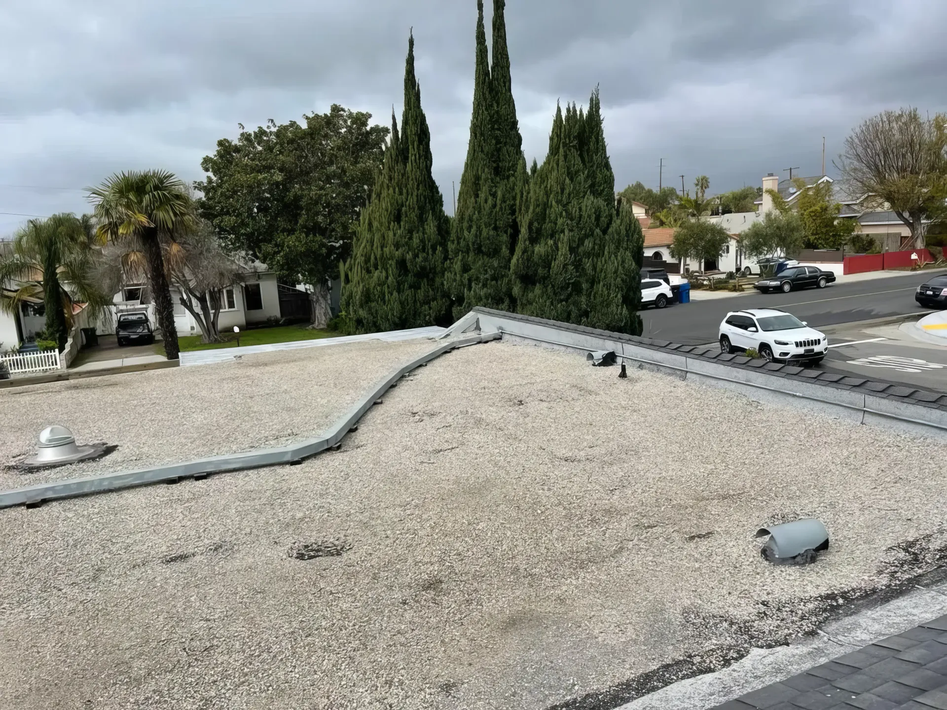 Gravel rooftop with vents and tall cypress trees beyond, overlooking a parking lot and street under cloudy skies