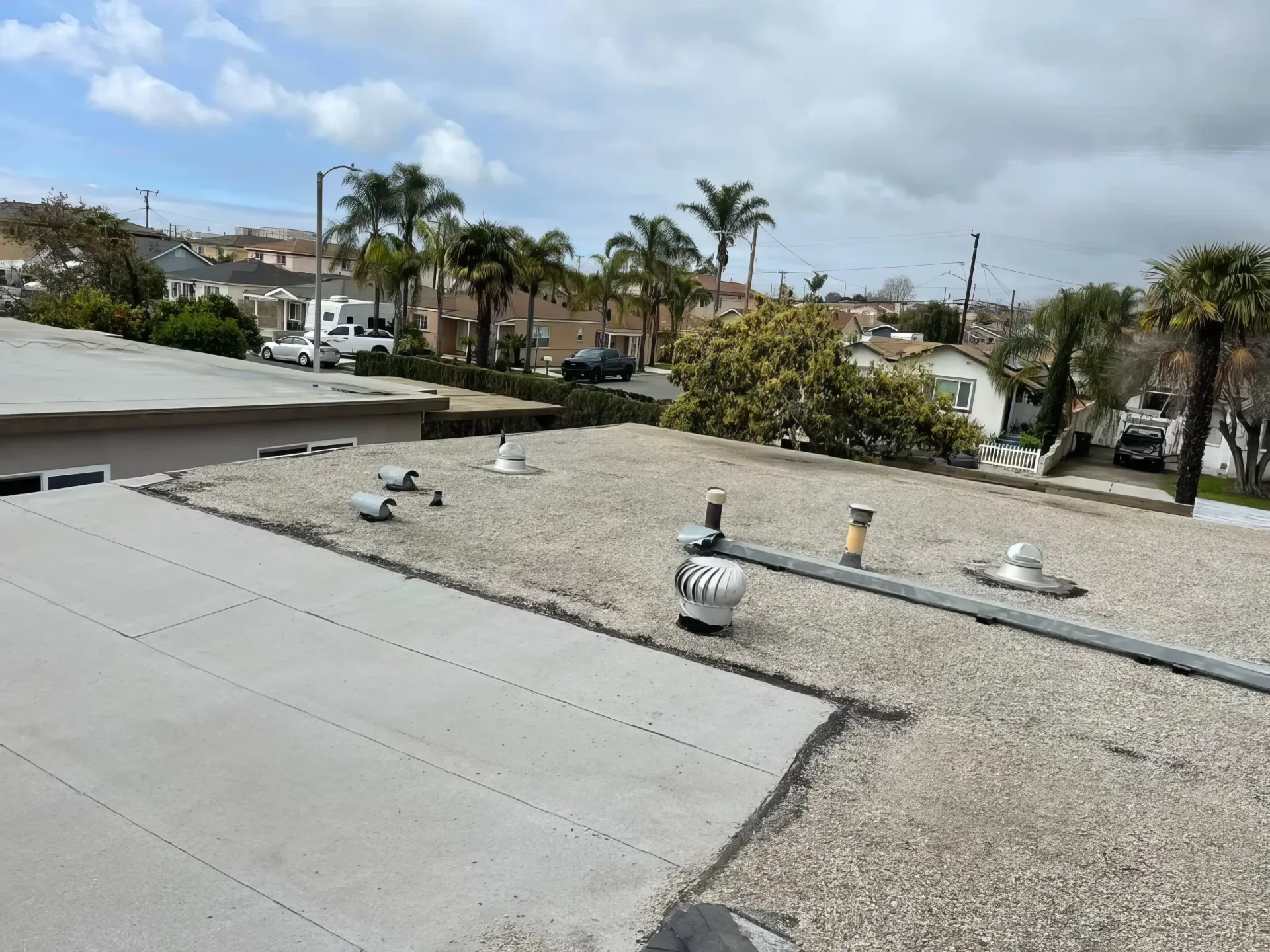 Rooftop with gravel and concrete surfaces, vents, and palm trees in the background under cloudy skies