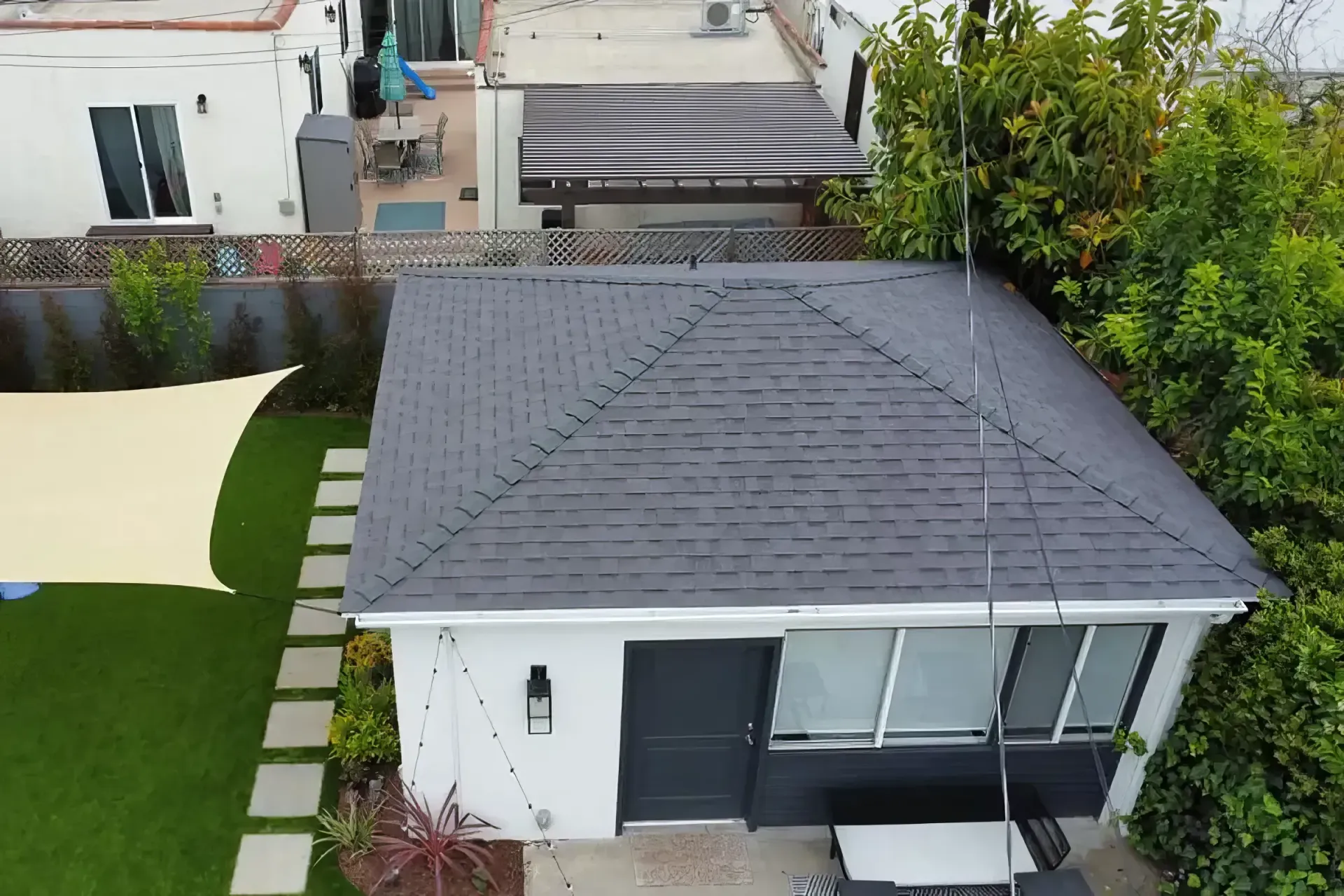 Aerial view of a small white house with a dark gray roof, patio, and green lawn beside it.