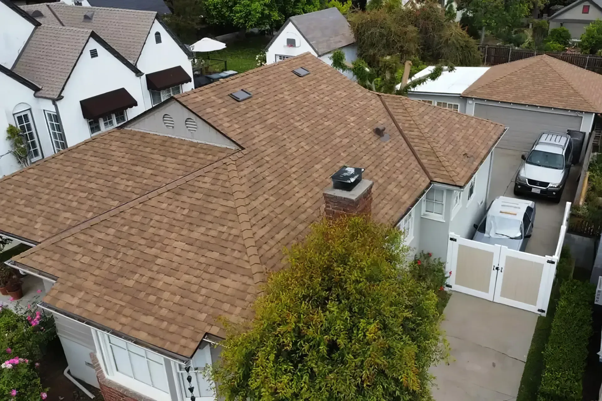 Aerial view of a suburban house with a tan roof, chimney, and neighboring homes along a driveway.