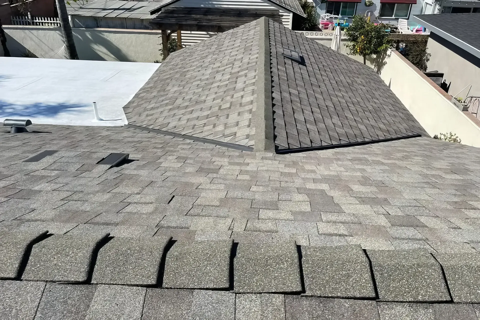 Roof shingles with a central ridge and nearby rooftops under bright daylight