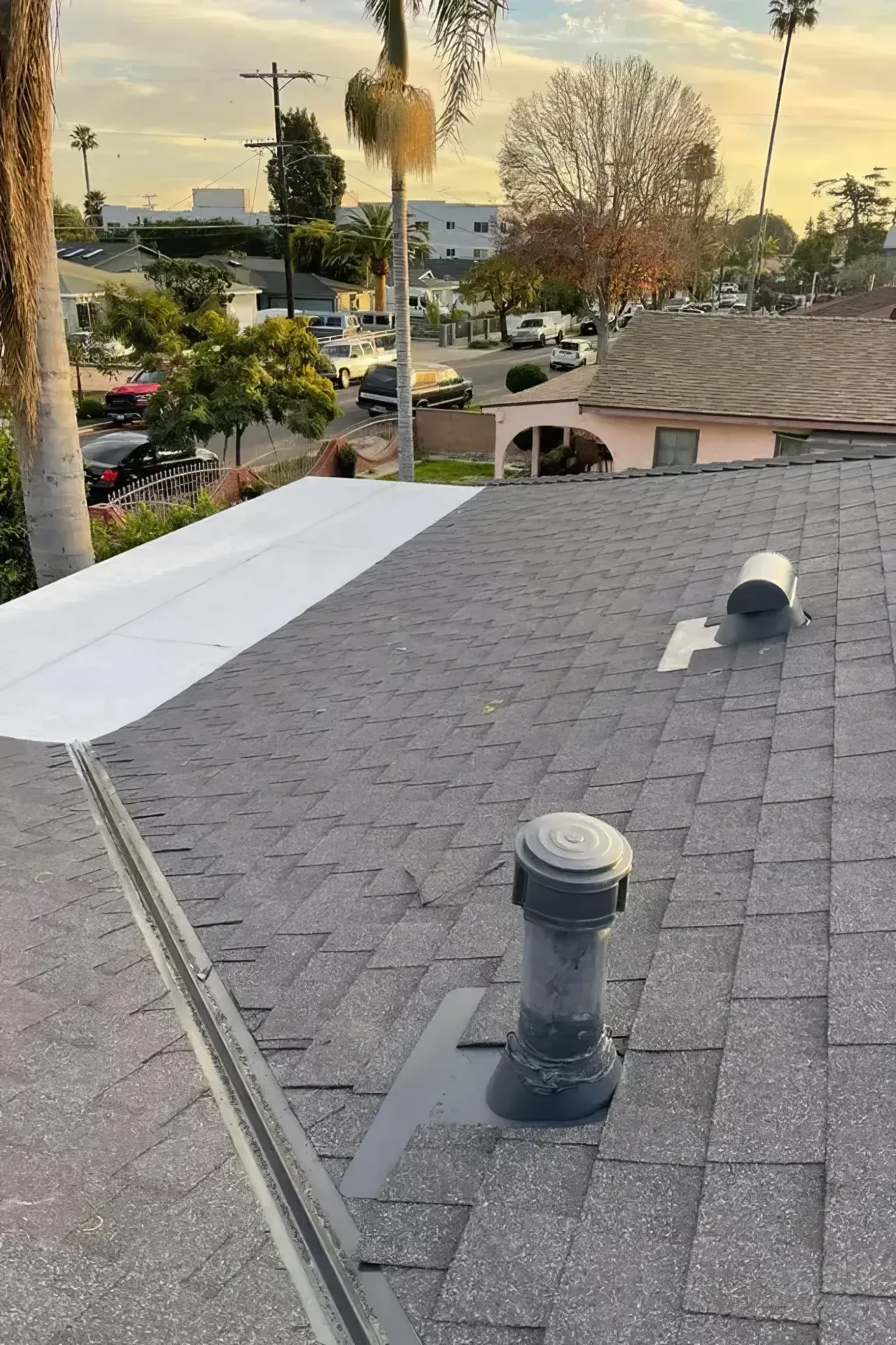 Rooftop view of gray shingles with vents, a white roof edge, and suburban homes at sunset