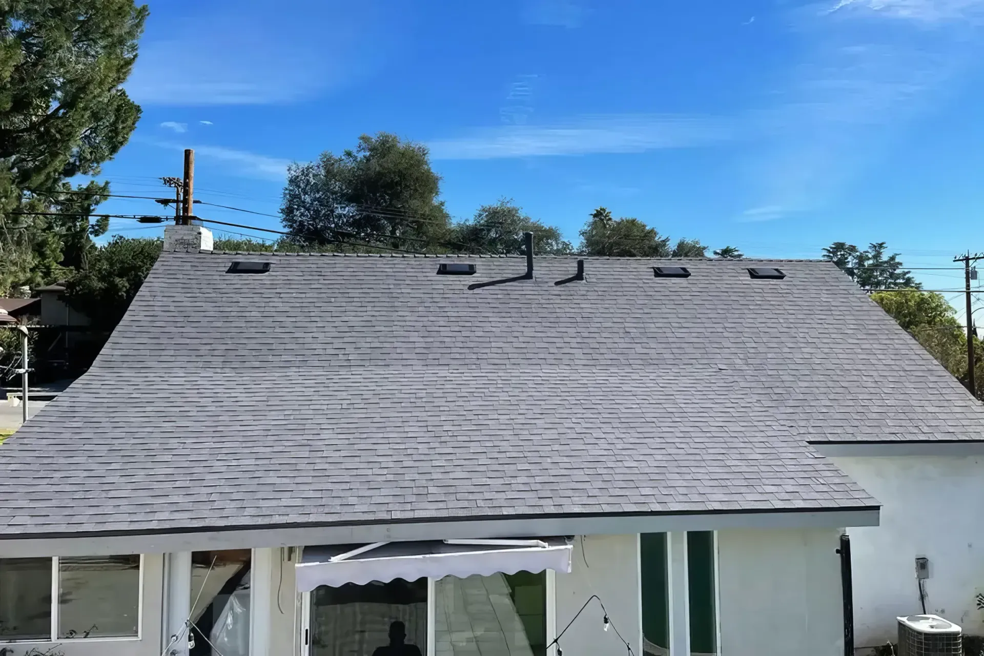 Gray shingled roof on a house under a clear blue sky, with trees and power lines in the background