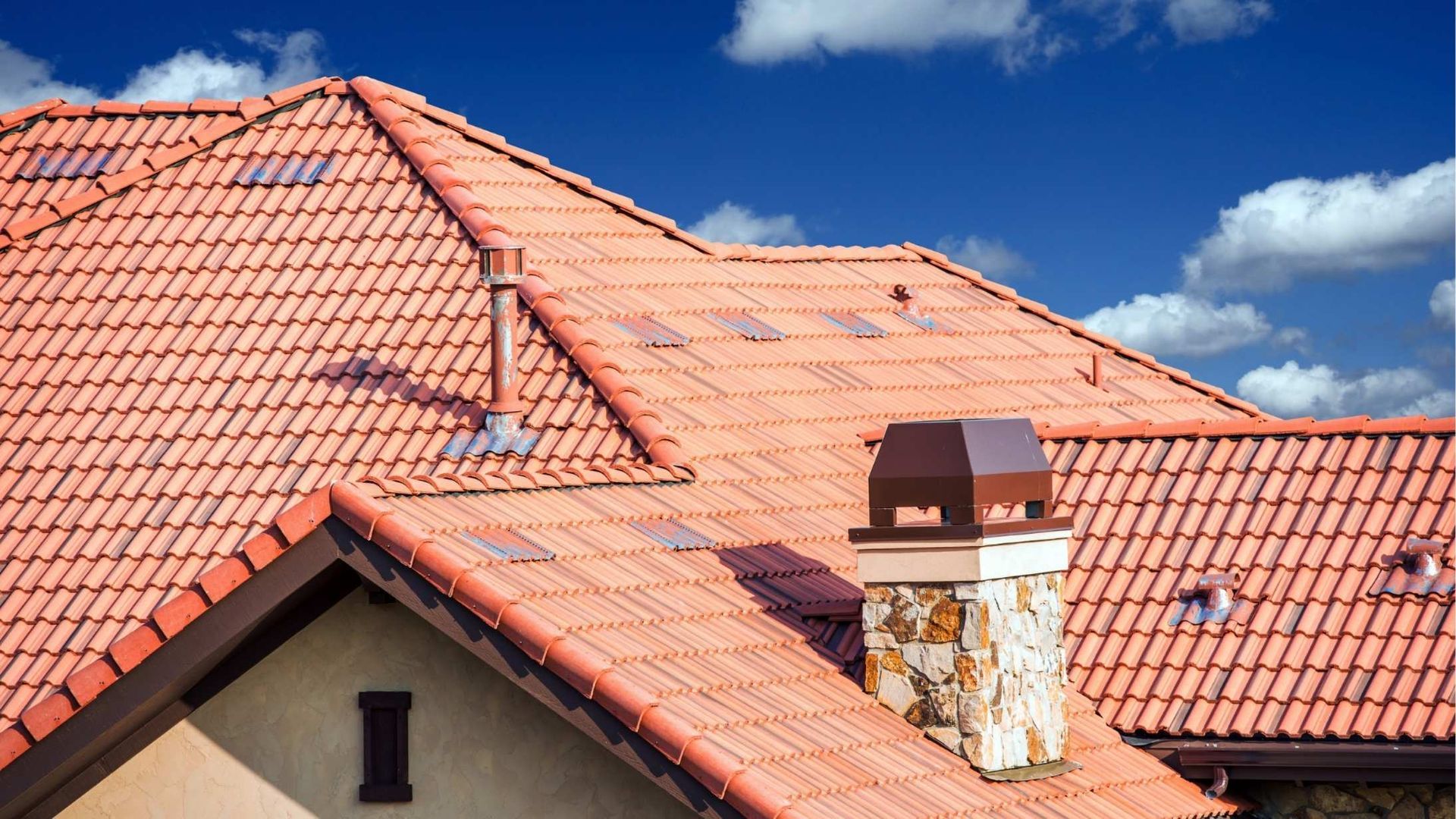 Red clay tile rooftop under blue sky with scattered clouds, featuring a chimney and dormer window.