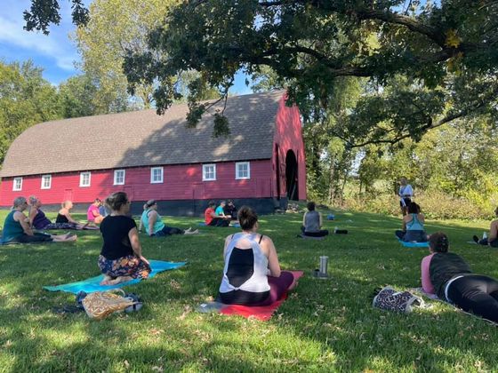 People doing yoga on mats in a sunny lawn beside a red barn under trees