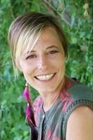 Smiling woman with short blonde hair standing outdoors against green foliage, wearing a colorful top