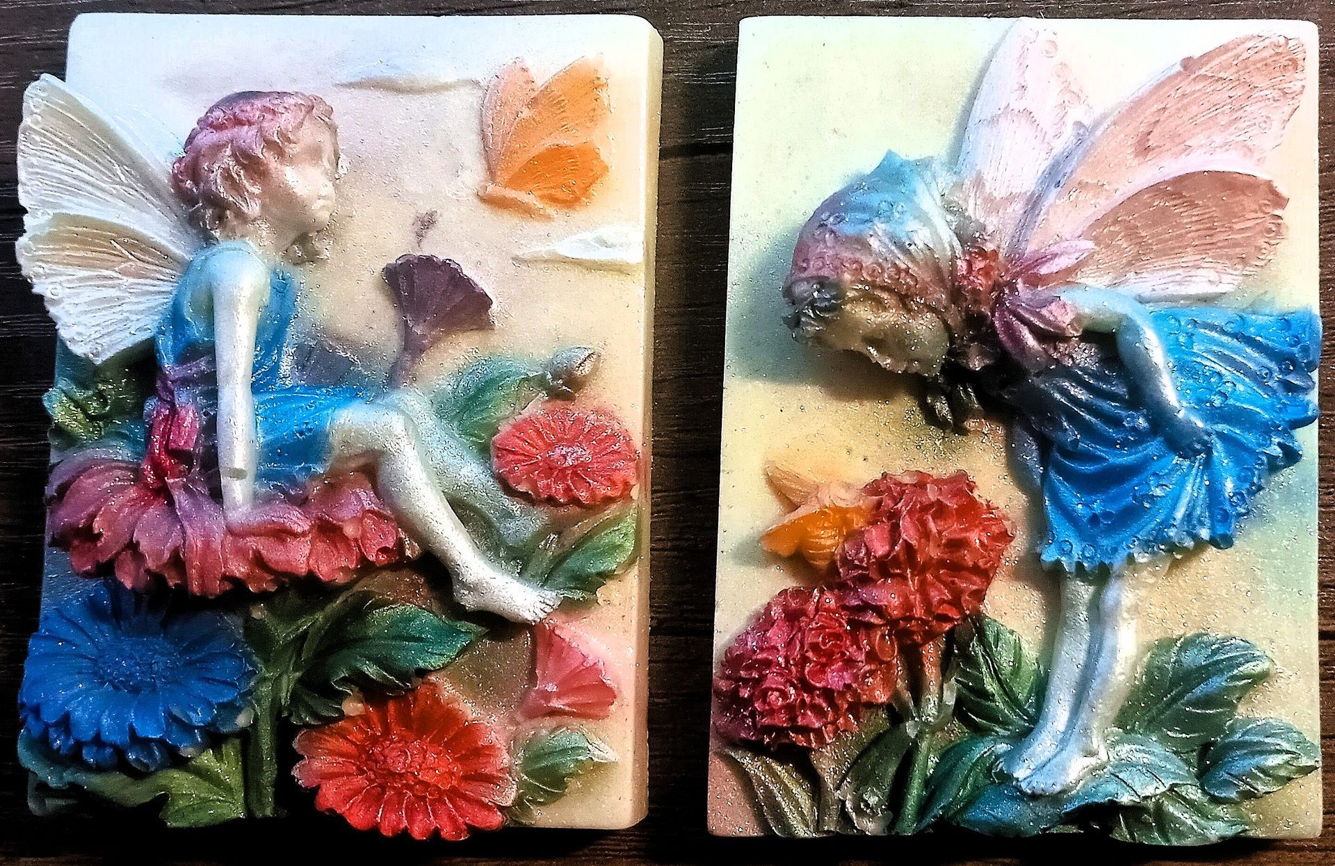 Two colorful  GamGams White Chocolate Art. Fairy figurines with blue, pink, and orange wings on a dark background