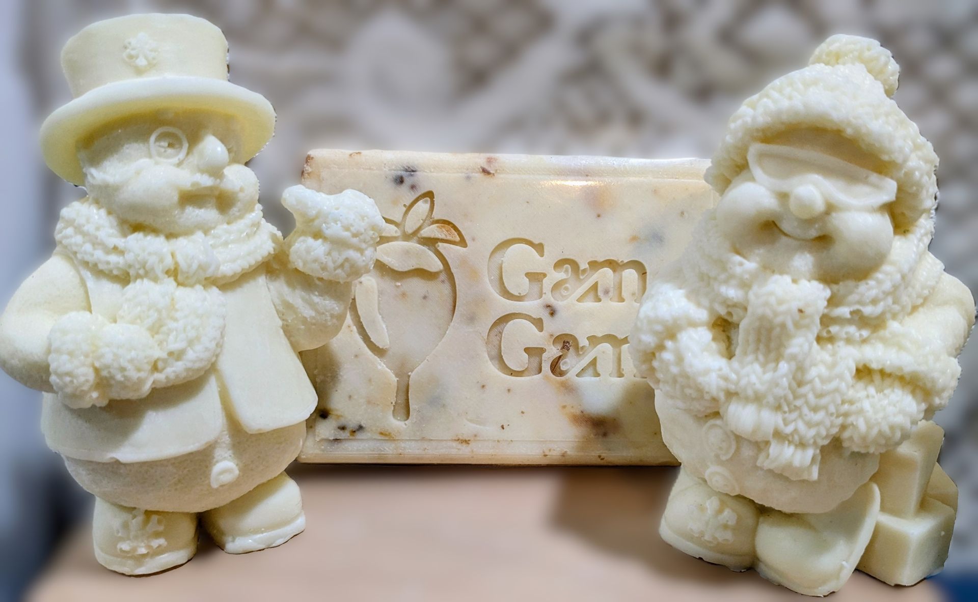 Two snowmen figurines flanking a white chocolate GamGams plaque with gold lettering and a rose design