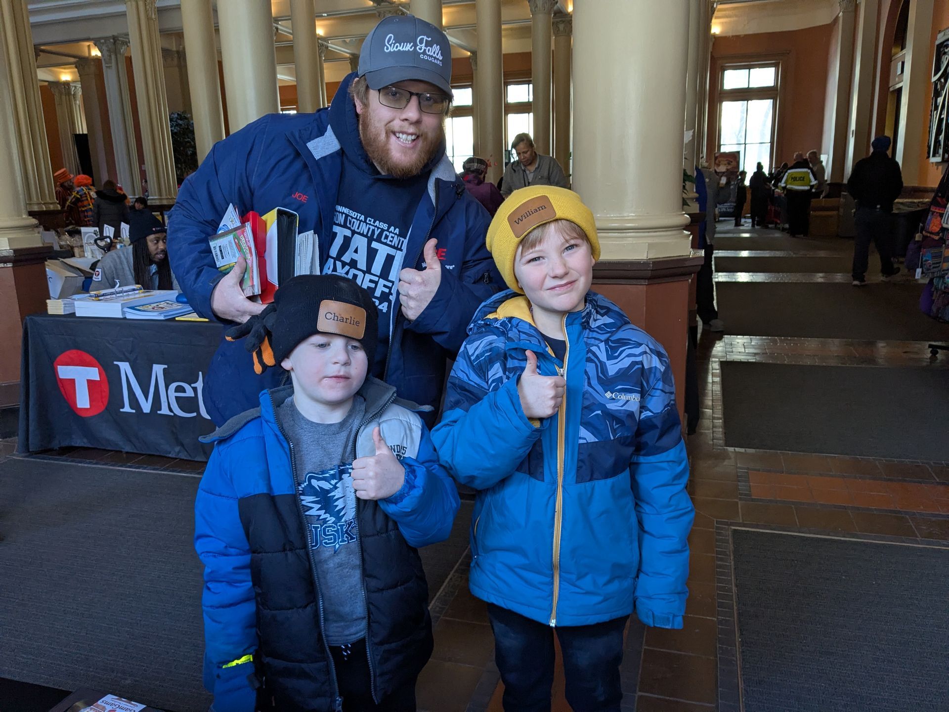Three people posing indoors, wearing blue winter jackets and hats, with thumbs up in the LandMark Center .