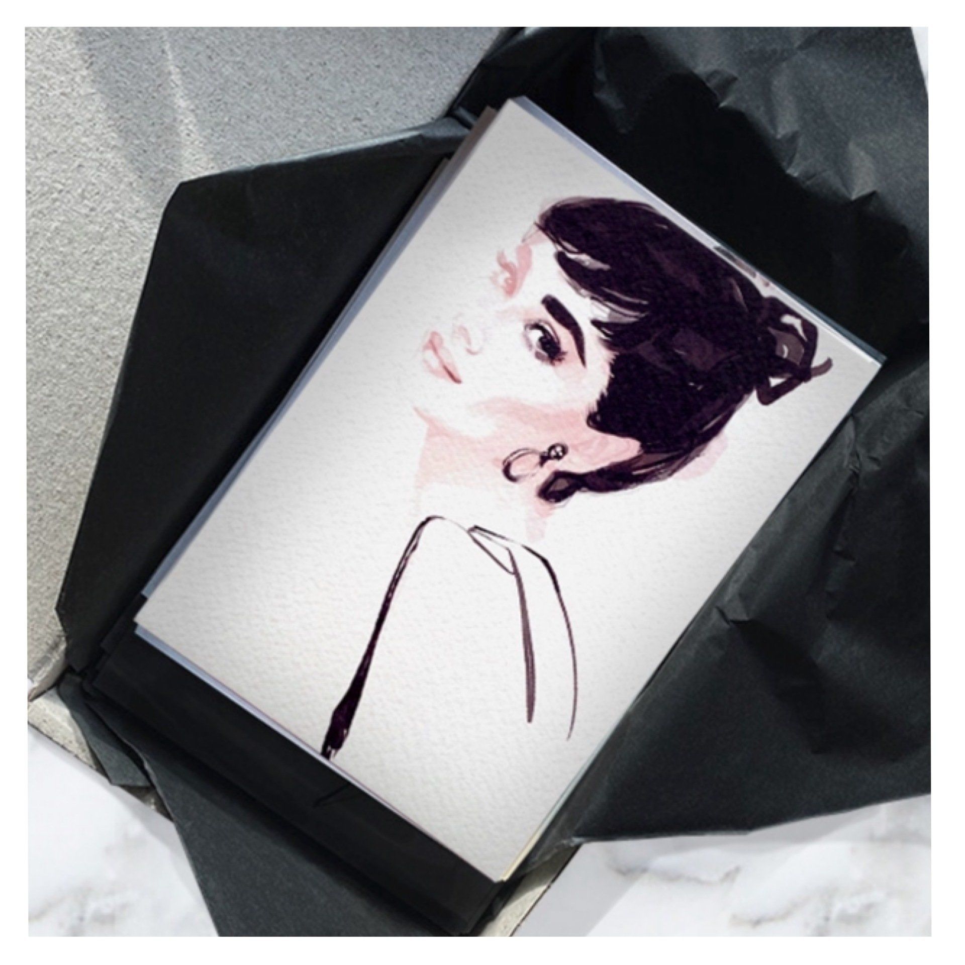 Watercolor portrait of Audrey Hepburn in a black bun, pale skin, simple black ink lines, in a black envelope.