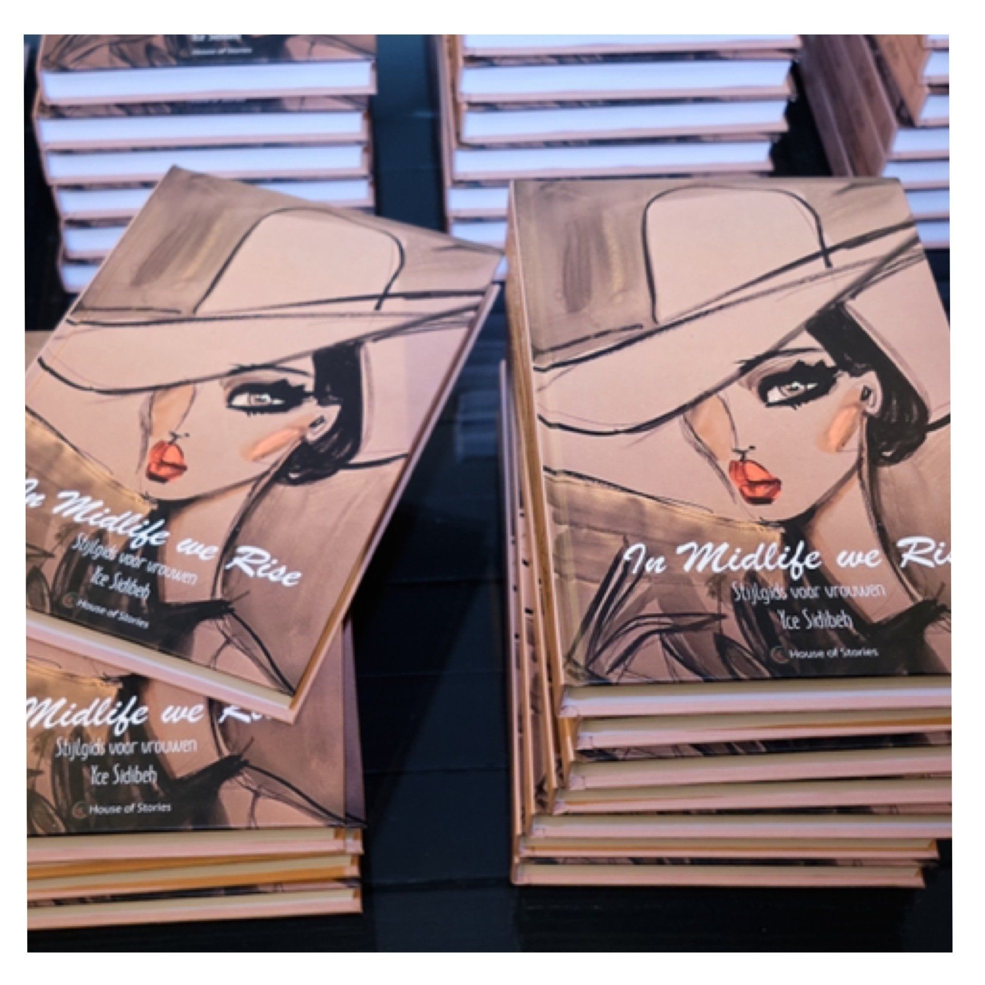 Stacks of books with cover art of a woman in a hat. The book title is βIn Midlife We Rise.β
