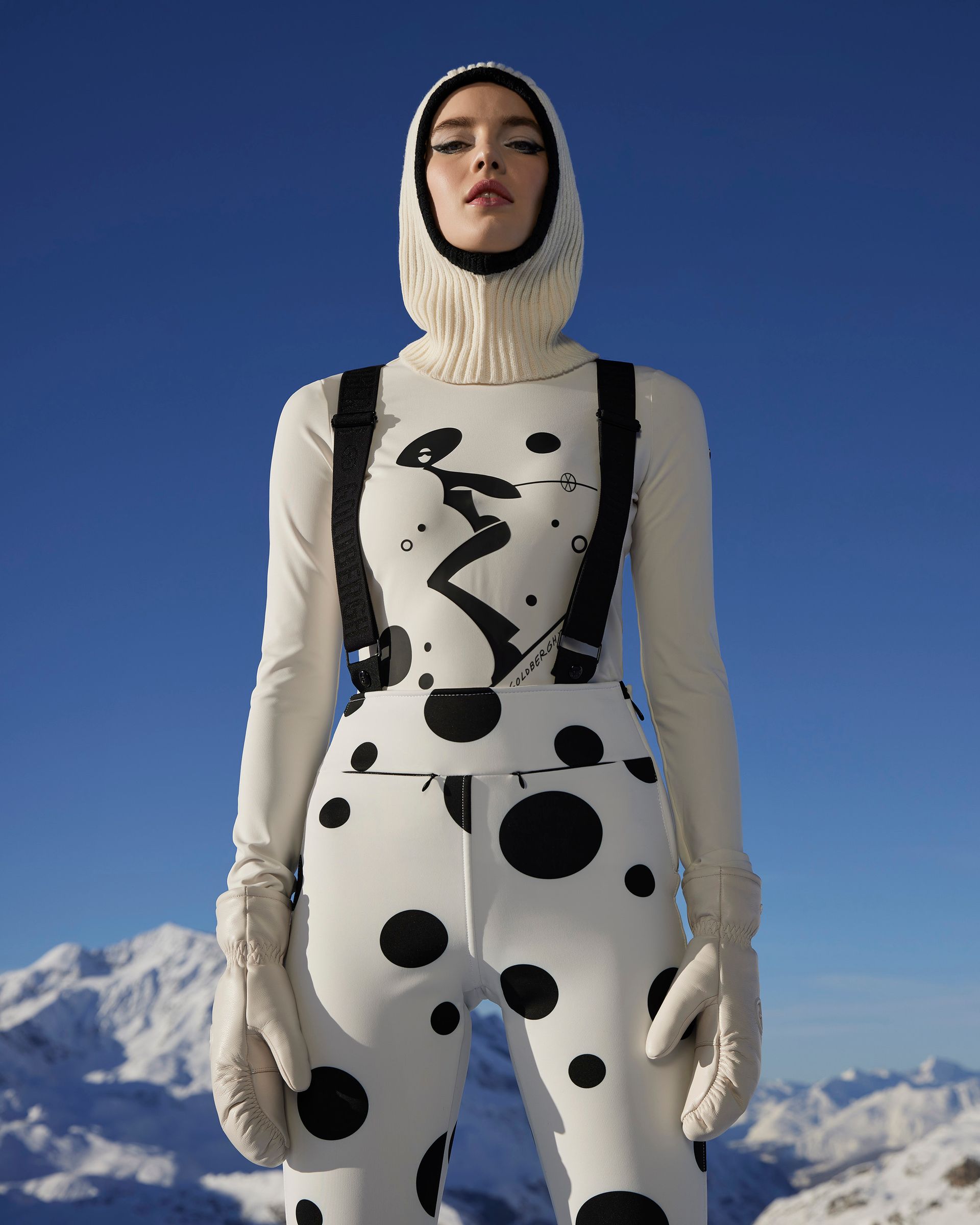 Woman in white and black polka-dot ski suit, standing on a snowy mountain.