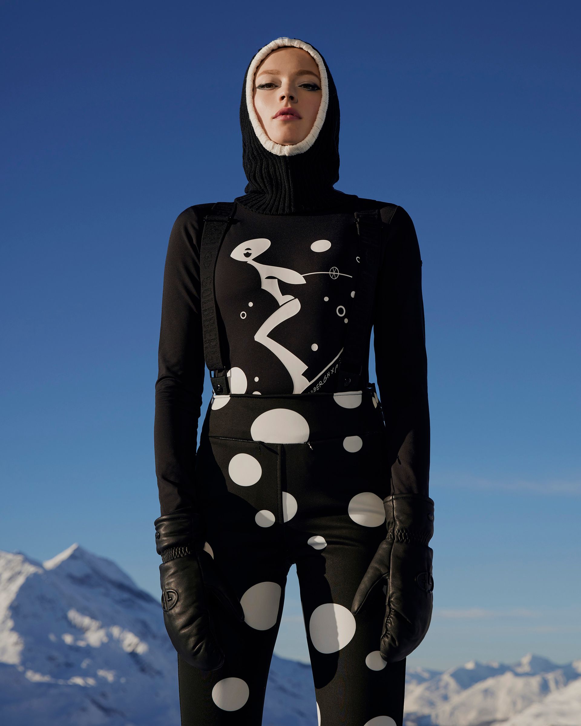 Woman in black and white ski suit and hood stands on a snowy mountain, blue sky backdrop.