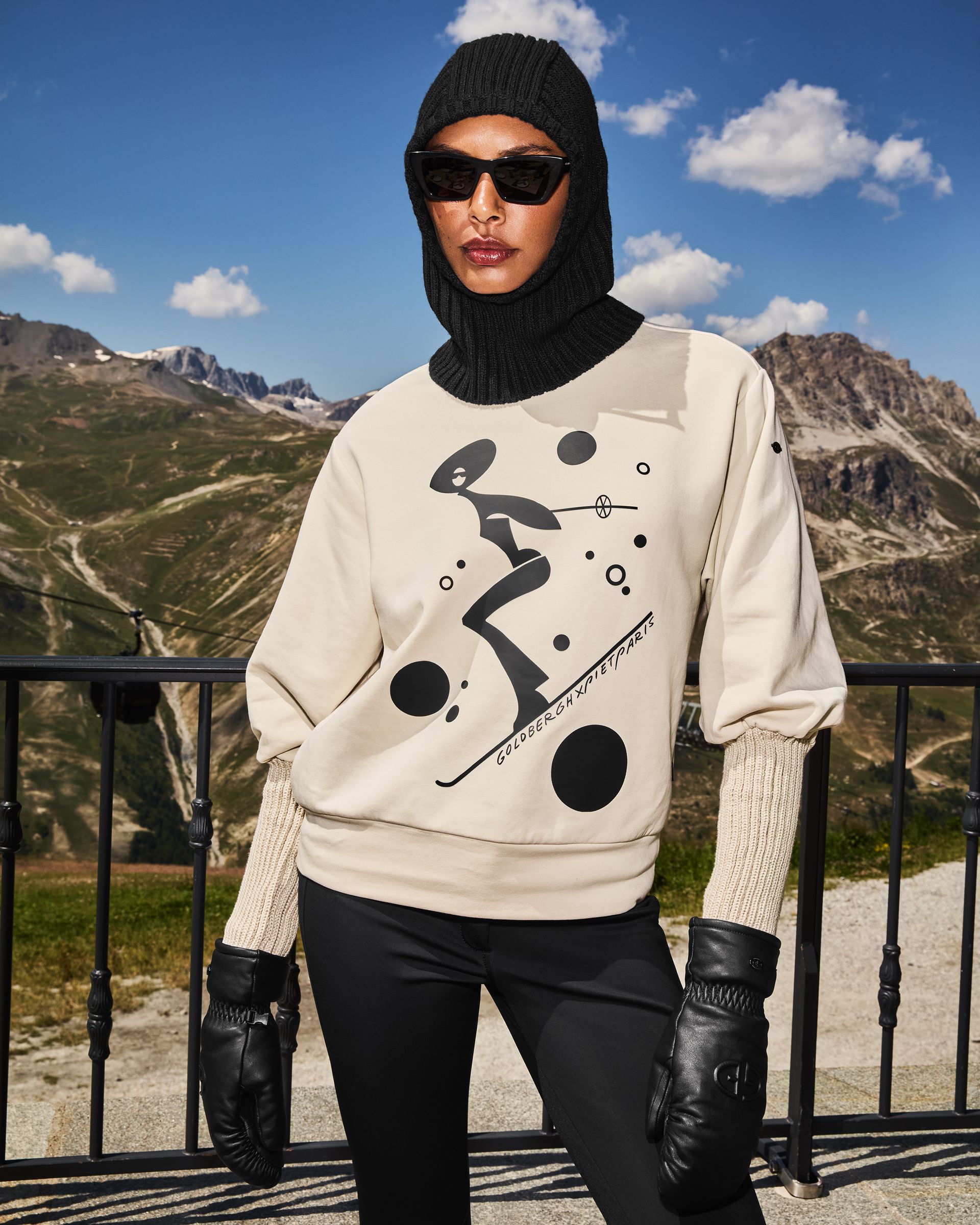 Person wearing black head covering, sunglasses, gloves, and a beige sweatshirt with black design; mountain backdrop.