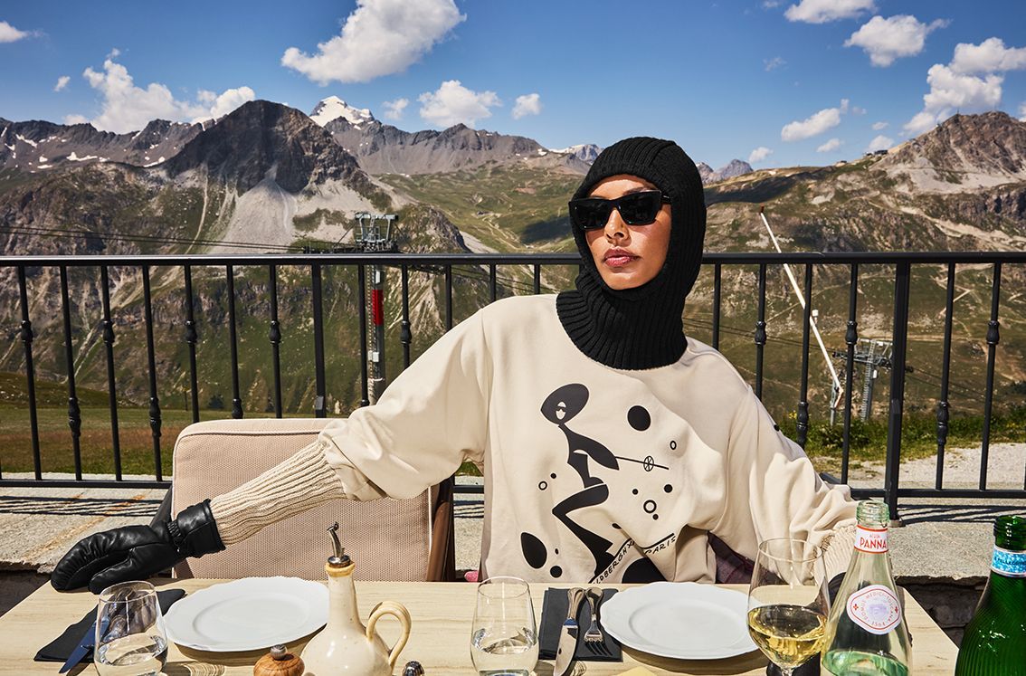 Person in black balaclava and sunglasses at an outdoor table with mountain view.