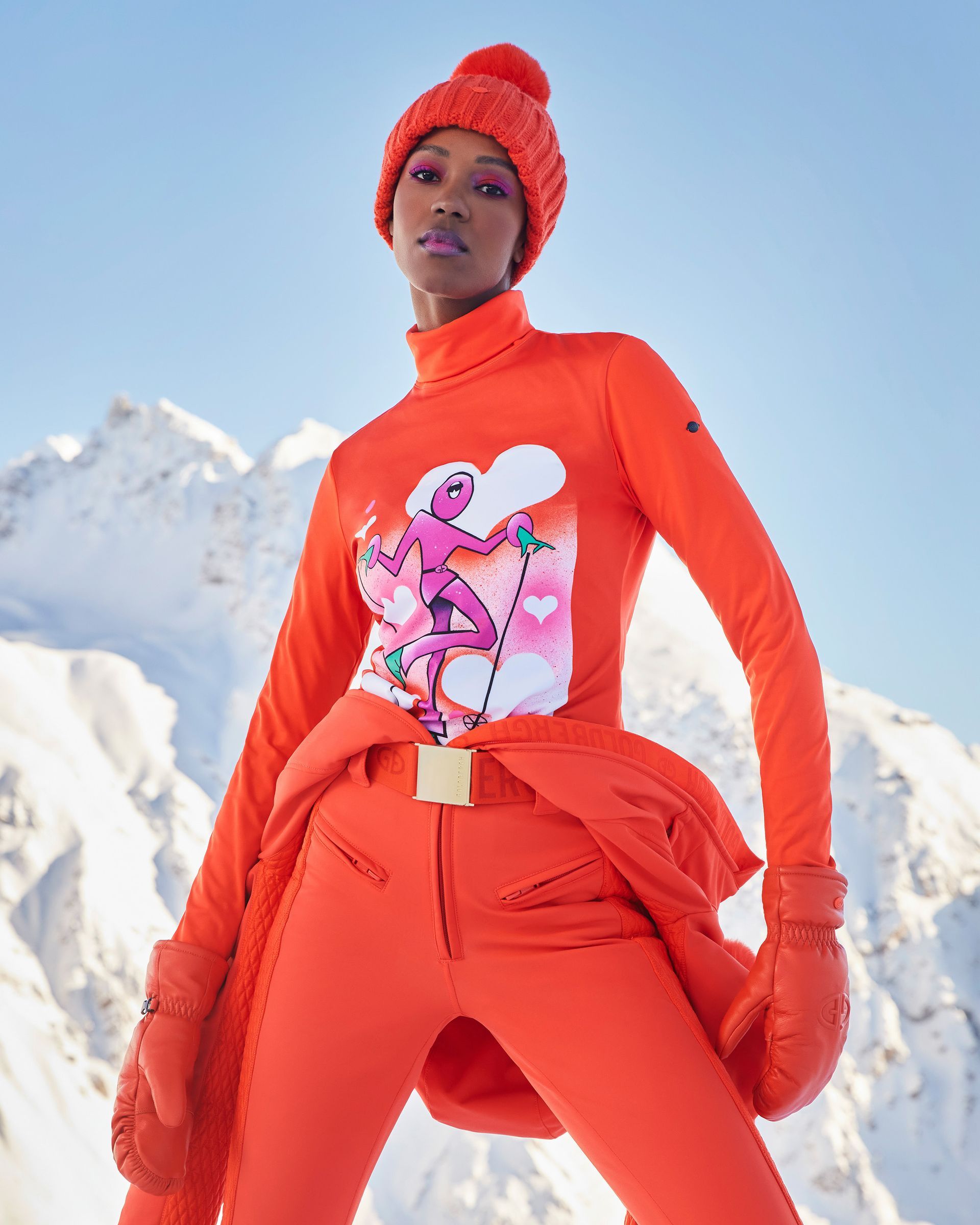Woman in orange ski outfit posing in front of snowy mountains; holding mittens.