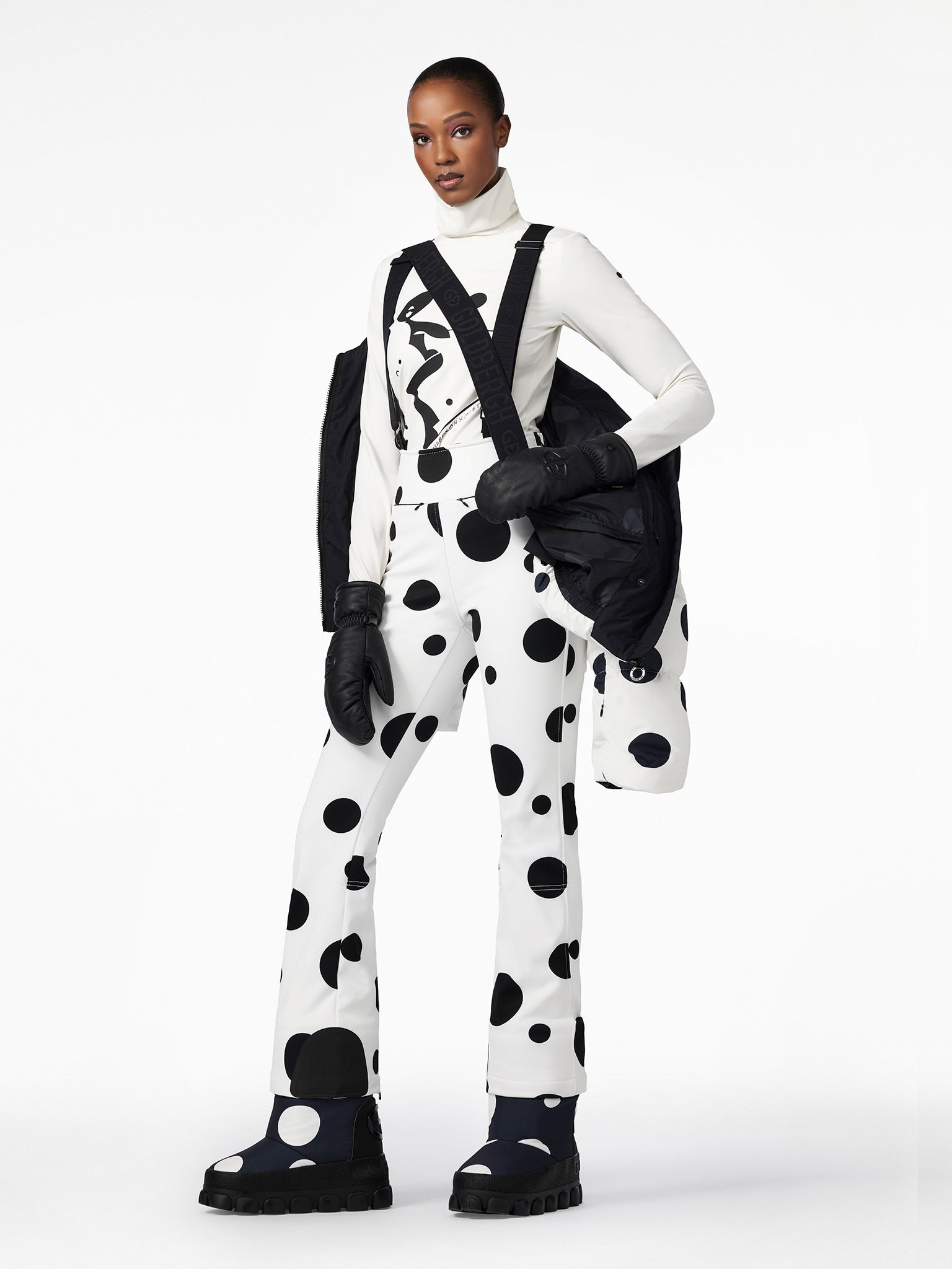 Woman in polka-dot jumpsuit and black jacket posing on white background.