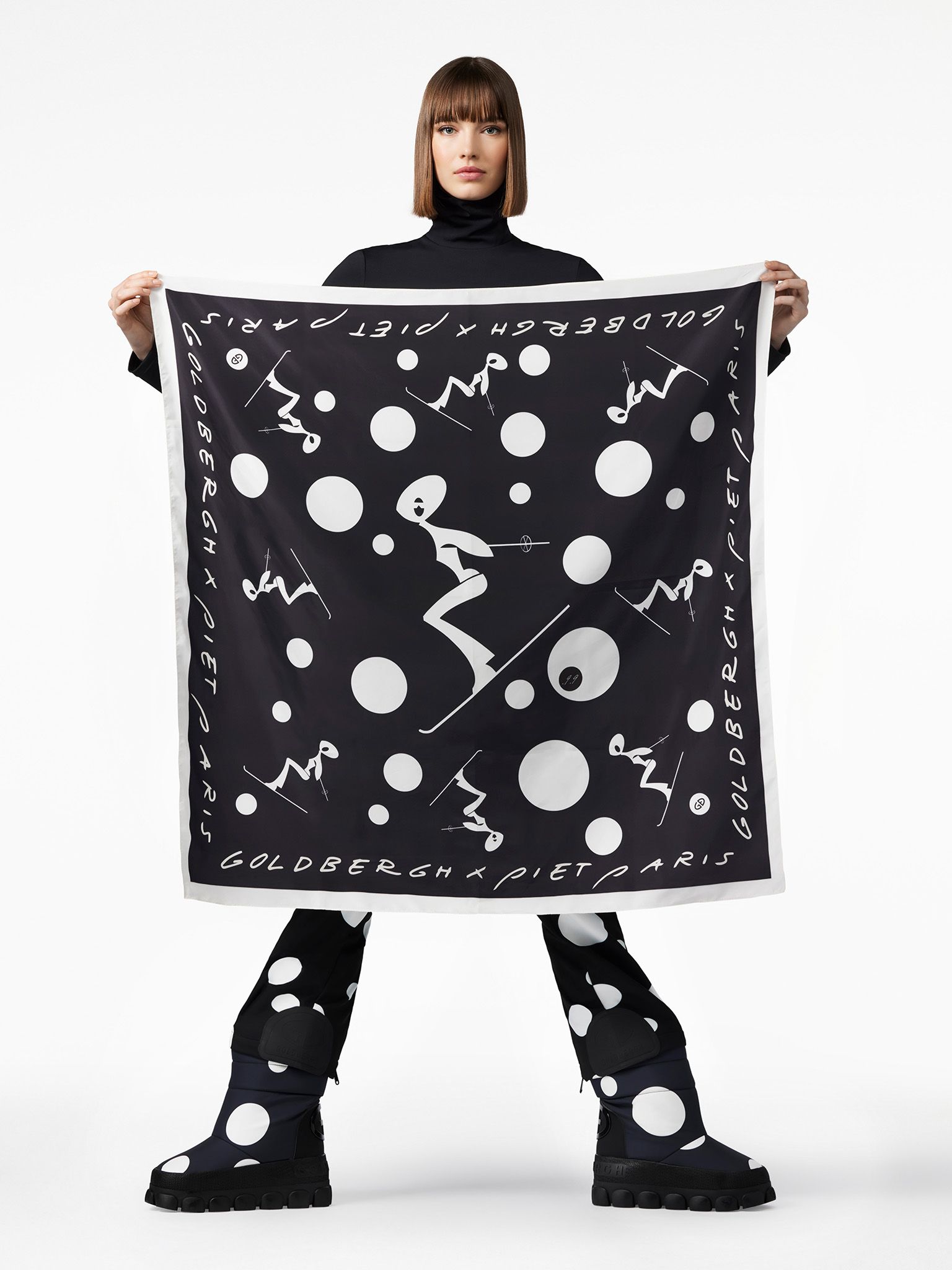 Woman holding a black and white patterned scarf. She wears a turtleneck and matching pants with large polka dots.
