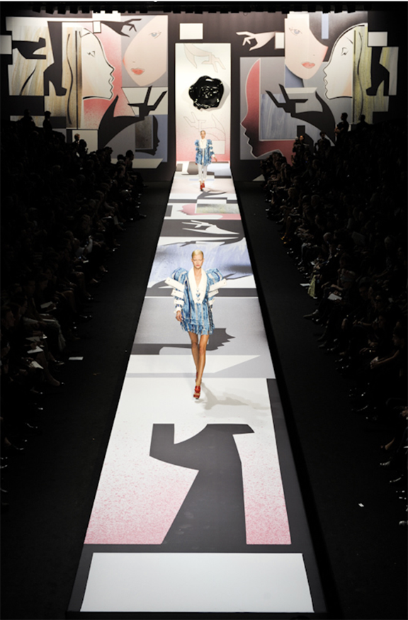 Fashion show runway with models in front of abstract art backdrop, bright lighting, and an audience.