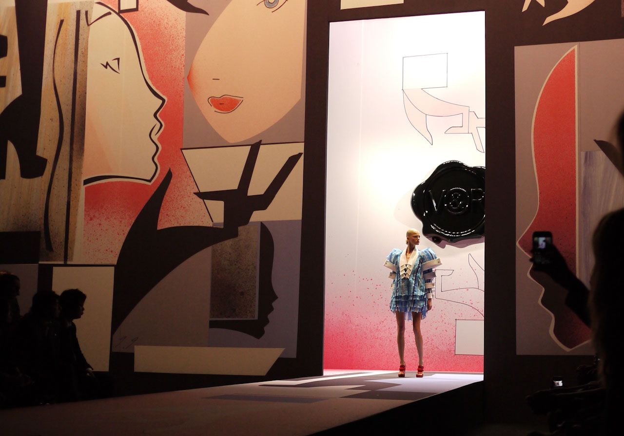 Fashion model on runway, wearing short dress. Colorful backdrop with face art. Spectators in shadows.