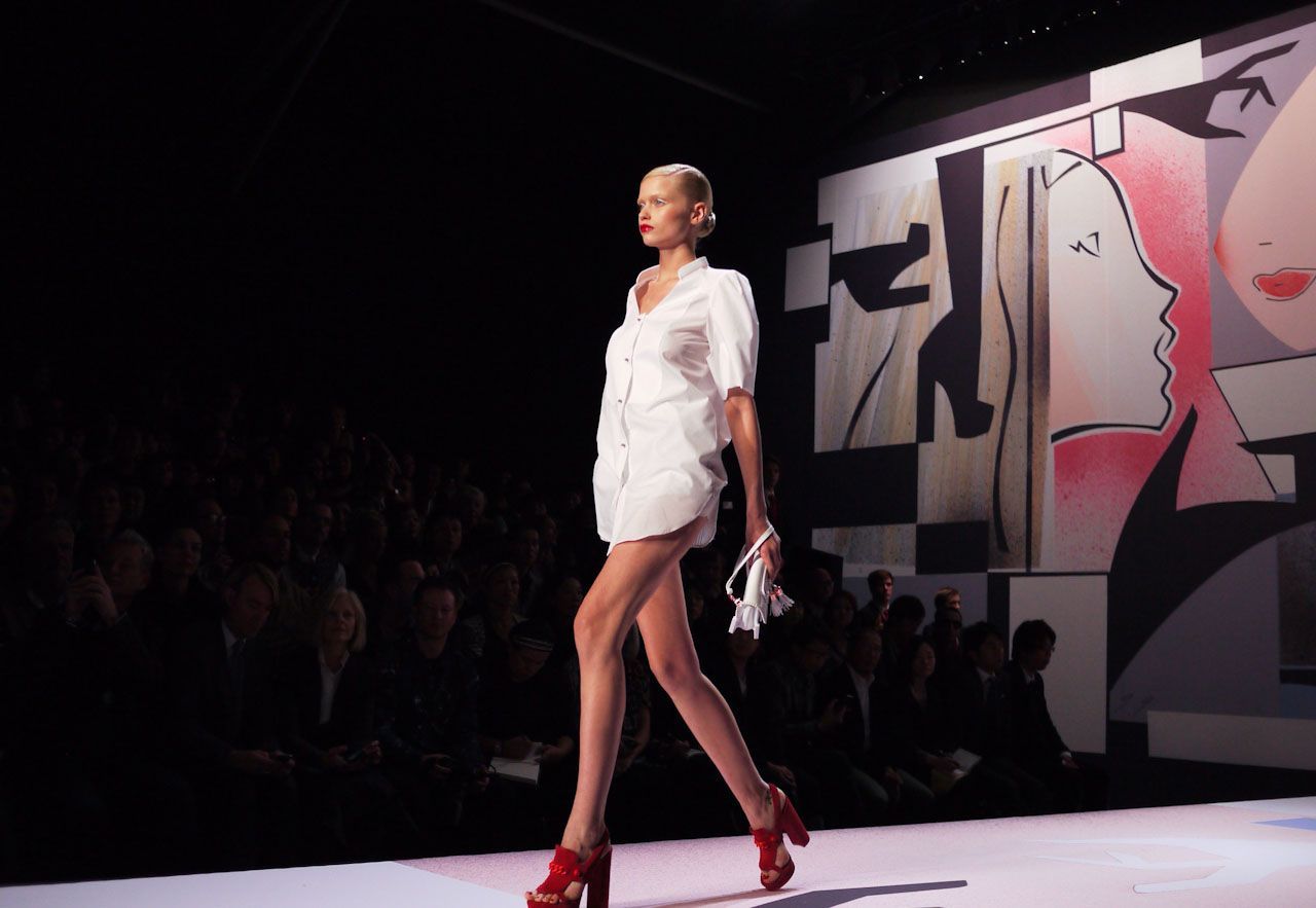 Model walks runway in white dress and red heels, carrying a white purse. Black background with audience.