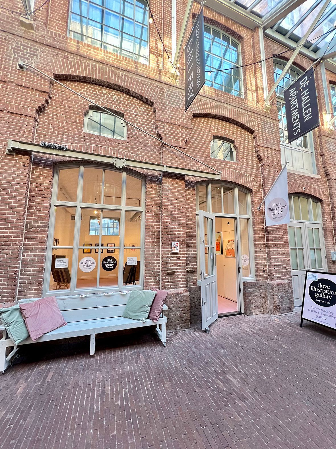 Brick building facade with glass windows and open doorway. A bench with pillows sits outside.Rental Space Venue at De Hallen Amsterdam