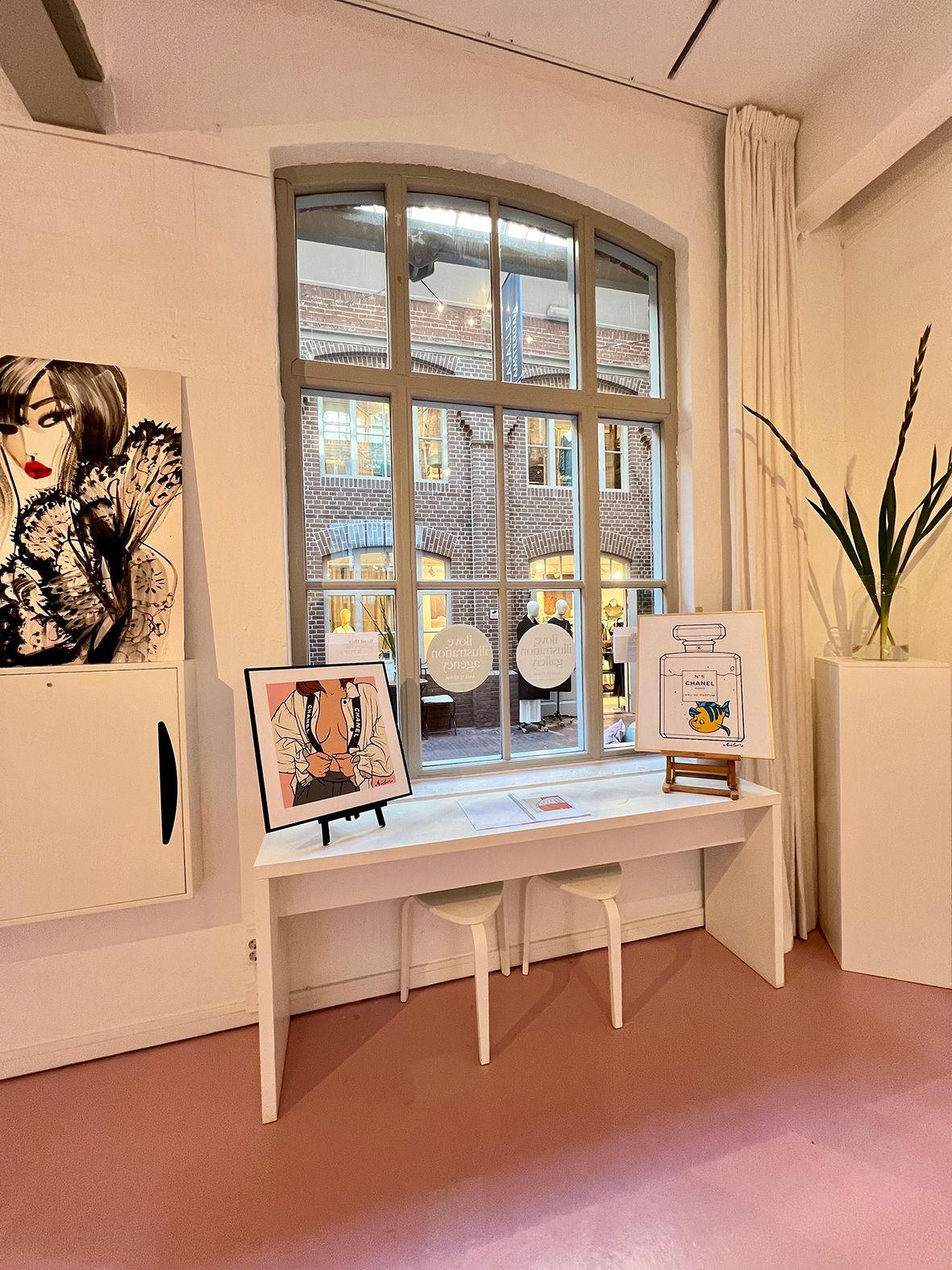 Art gallery display, white table with art prints, large arched window, exterior brick building view, pink carpet. Rental Space Venue at De Hallen Amsterdam