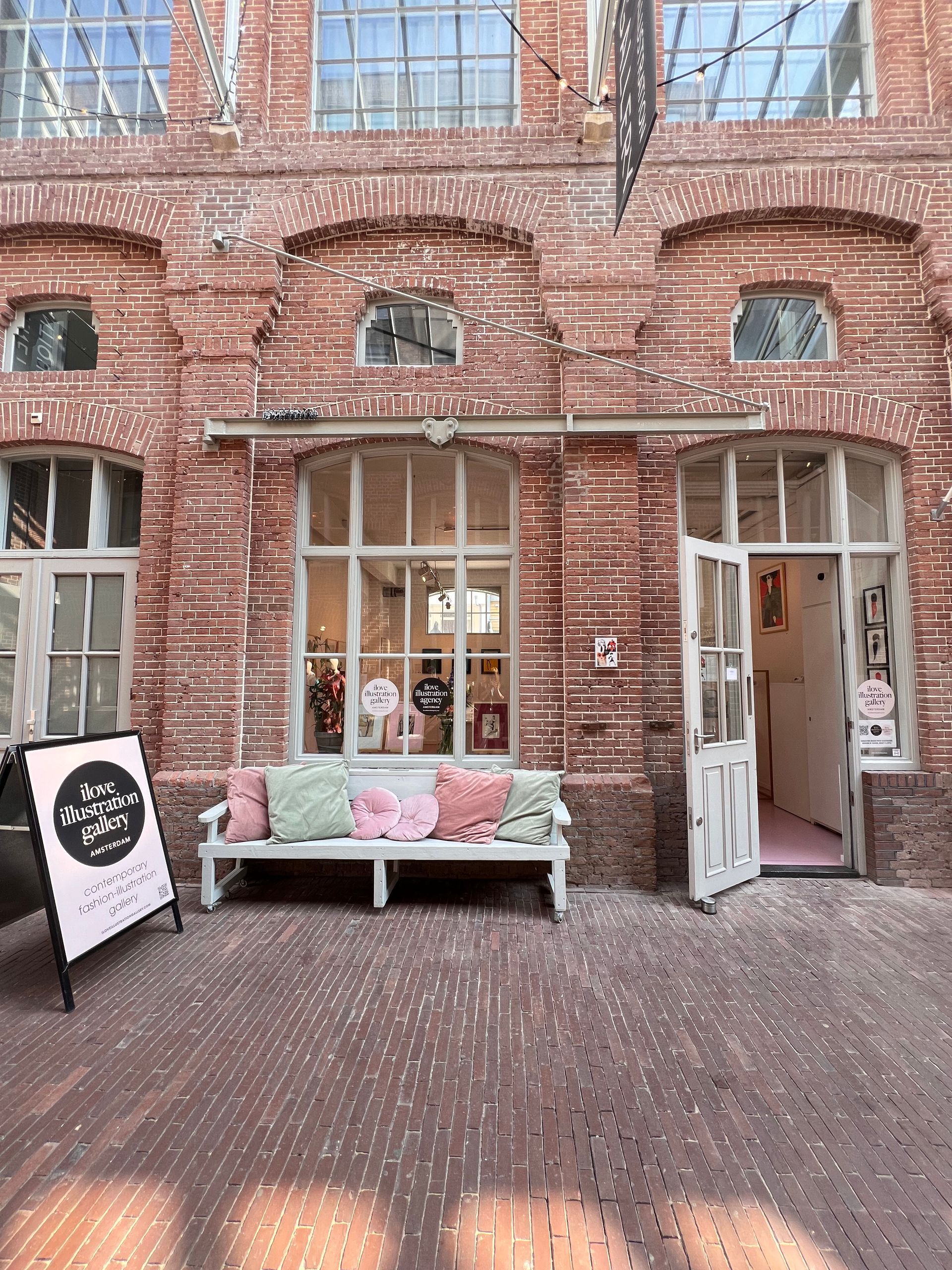 Brick building with shop entrance, white door ajar, bench with pillows. Sign on the left.Rental Space Venue at De Hallen Amsterdam