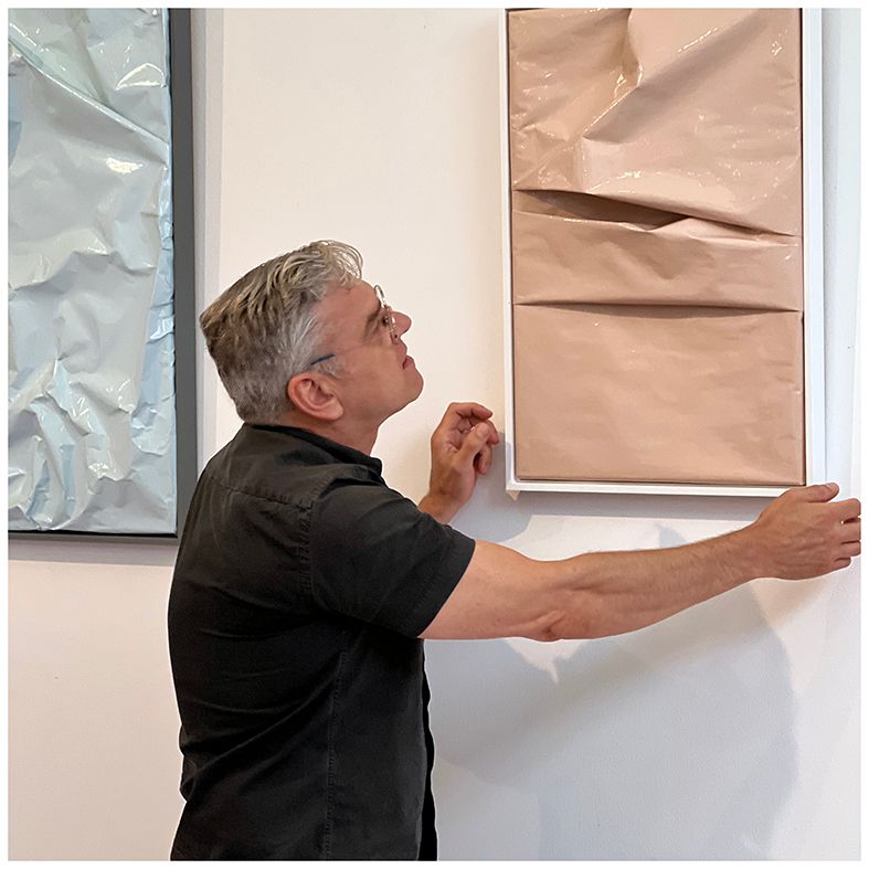 Man in black shirt examines abstract art piece on a white wall; tan paper with folds and white frame.