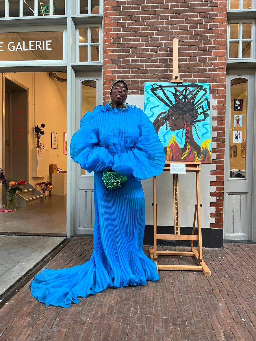 Person in a vibrant blue dress poses by their colorful artwork outside a gallery.