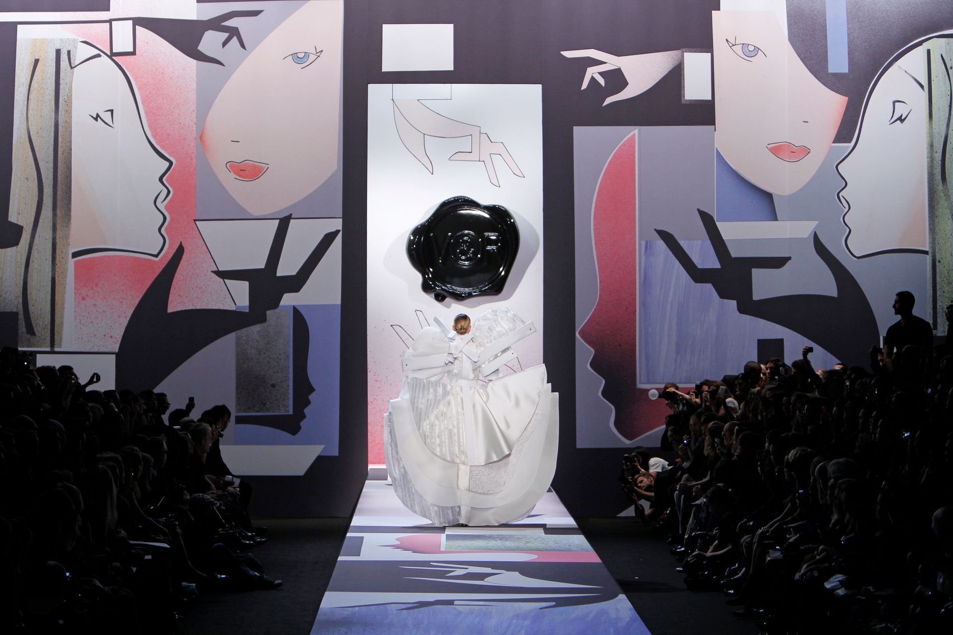 Fashion show runway. Model in white dress with large black embellishment. Backdrop of stylized faces in pastel colors.