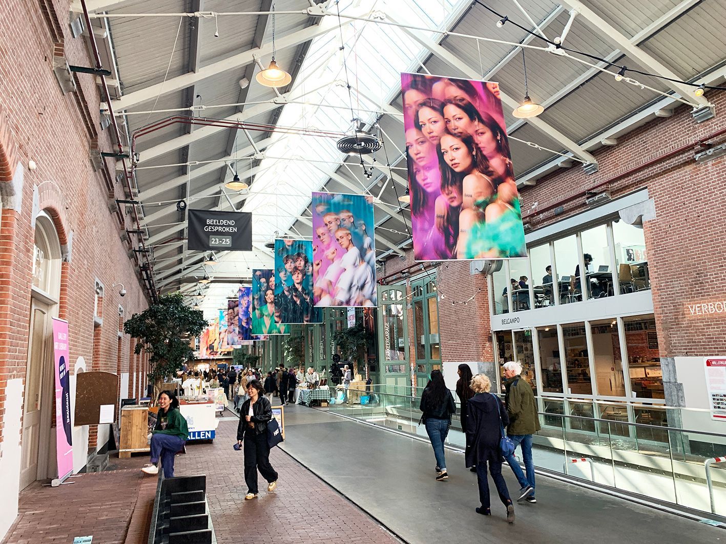 People walking through a brightly lit indoor marketplace with large colorful banners hanging from the ceiling.Rental Space Venue at De Hallen Amsterdam