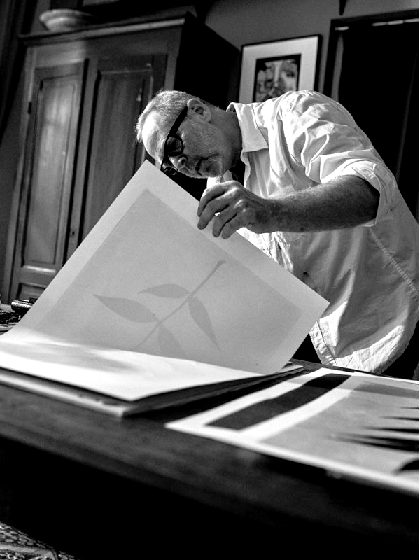 Man examining print, bending over table; light from window, monochromatic photo.
