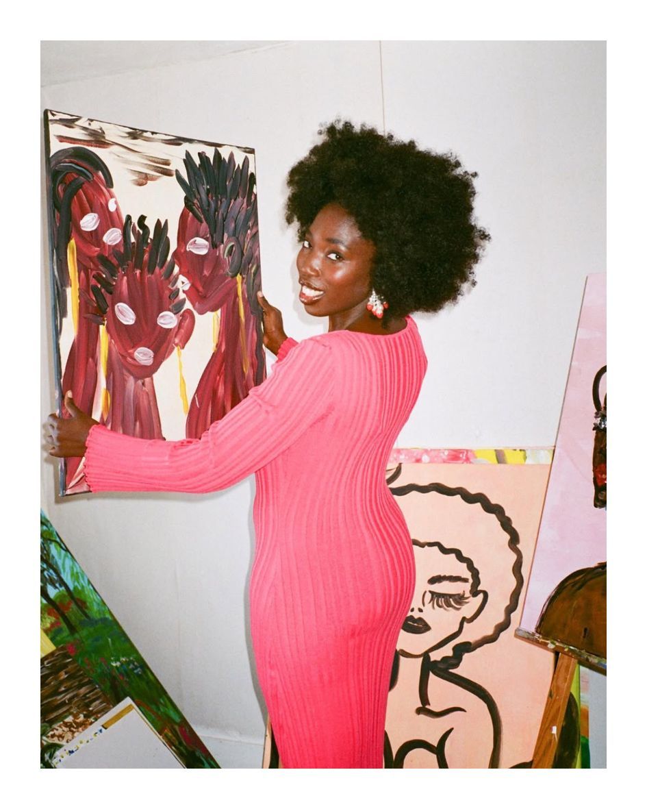 Woman in pink dress hanging art; curly hair, smiling, white wall background, several paintings.