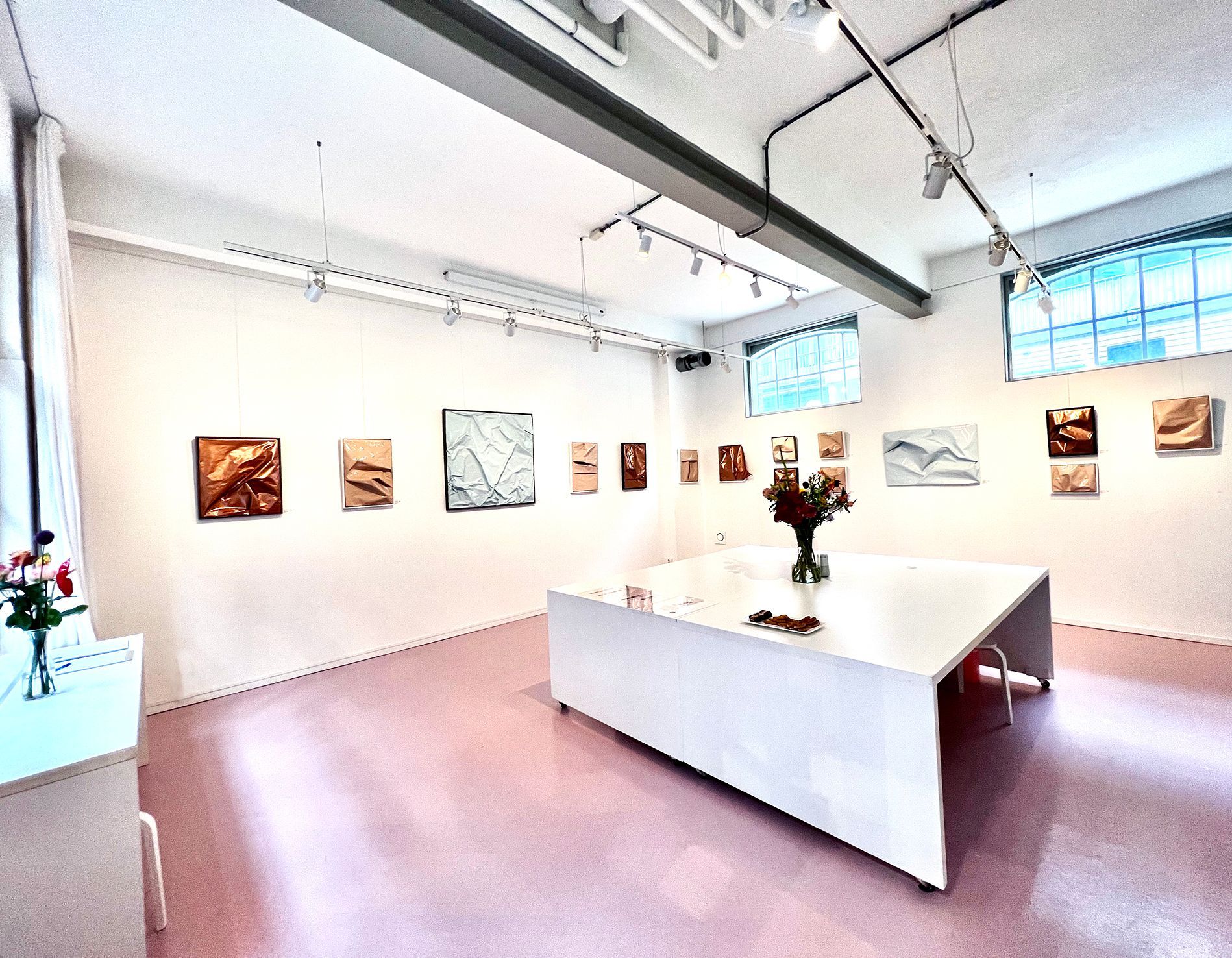 Art gallery with paintings on white walls, pink floor, large white angled table, and flower arrangement.Rental Space Venue at De Hallen Amsterdam