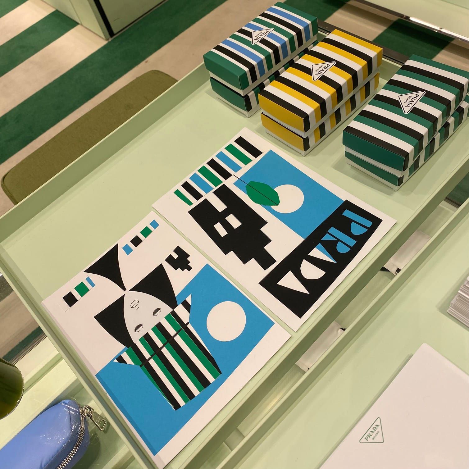 Prada display: striped boxes and art prints in a light green tray; green, blue, yellow, black, and white colors.
