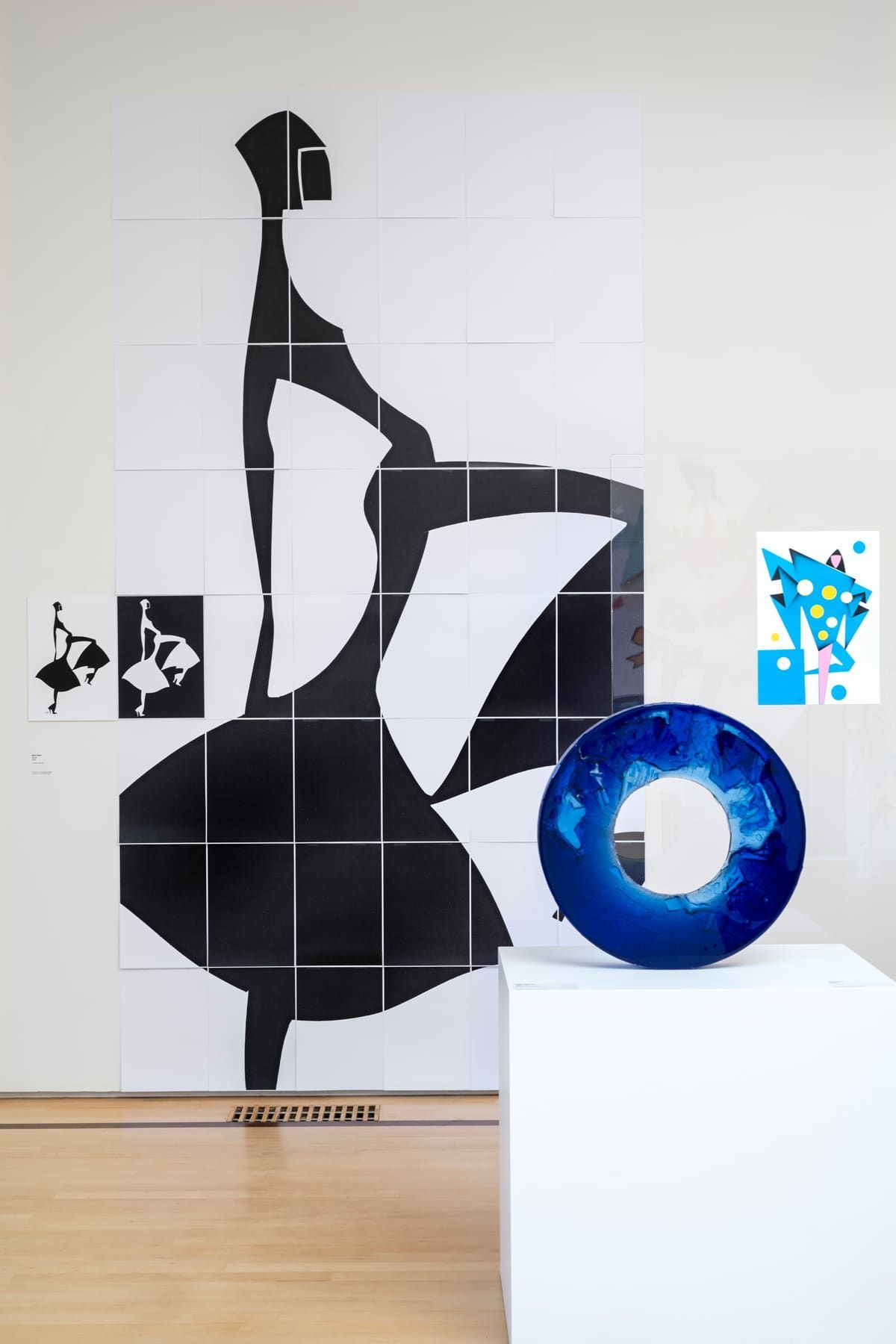 Black silhouette of a dancer on a wall composed of tiles, with a blue glass sculpture and white pedestal in the foreground.