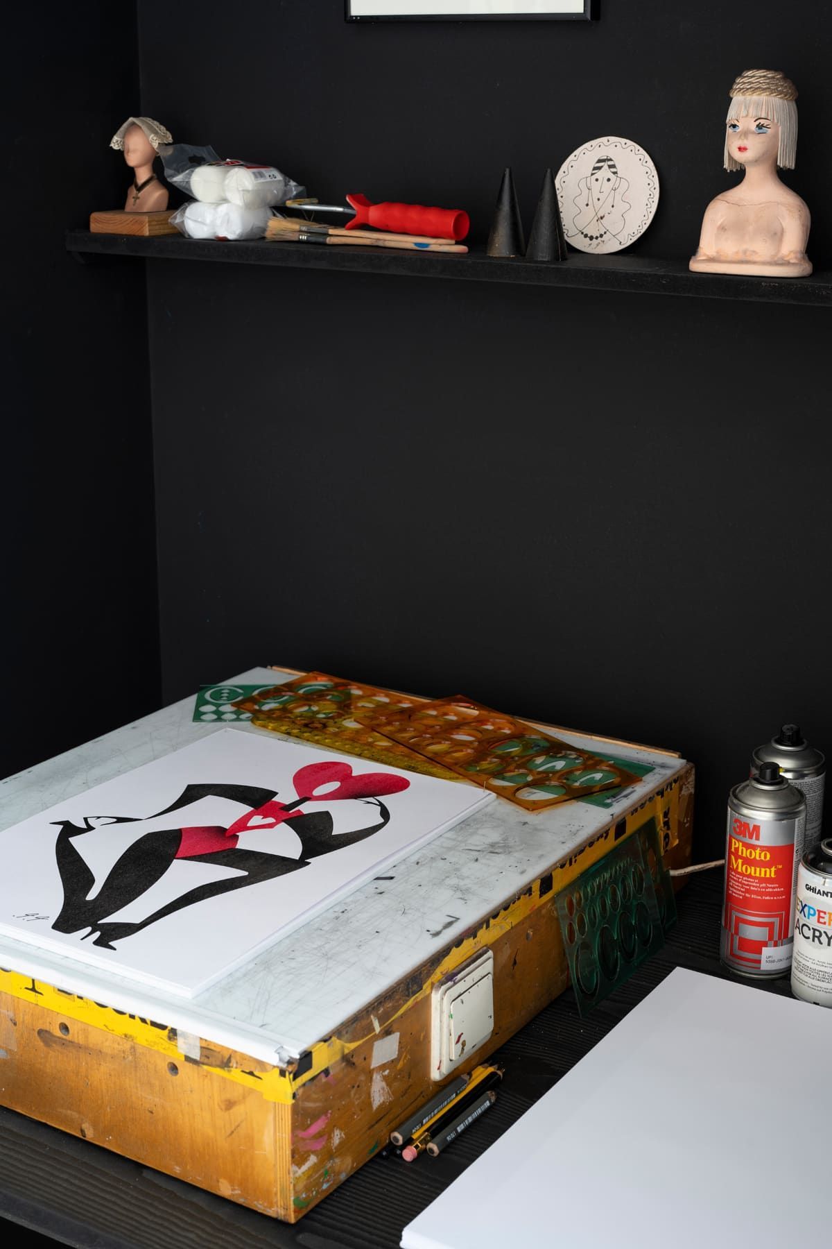 Workspace with artwork on a light table, shelf with objects, and black walls.