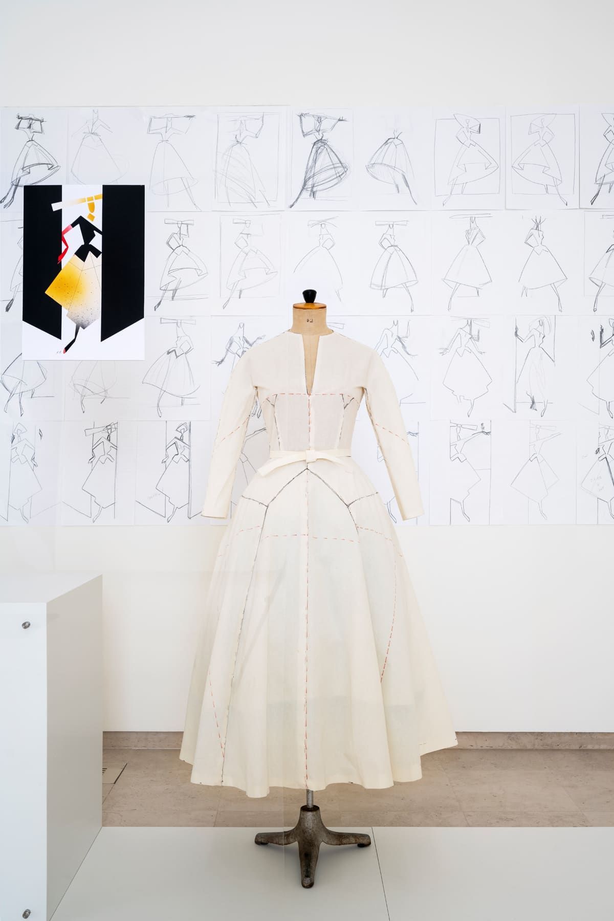 A white dress form with a white dress on a stand, in front of a wall with drawings.