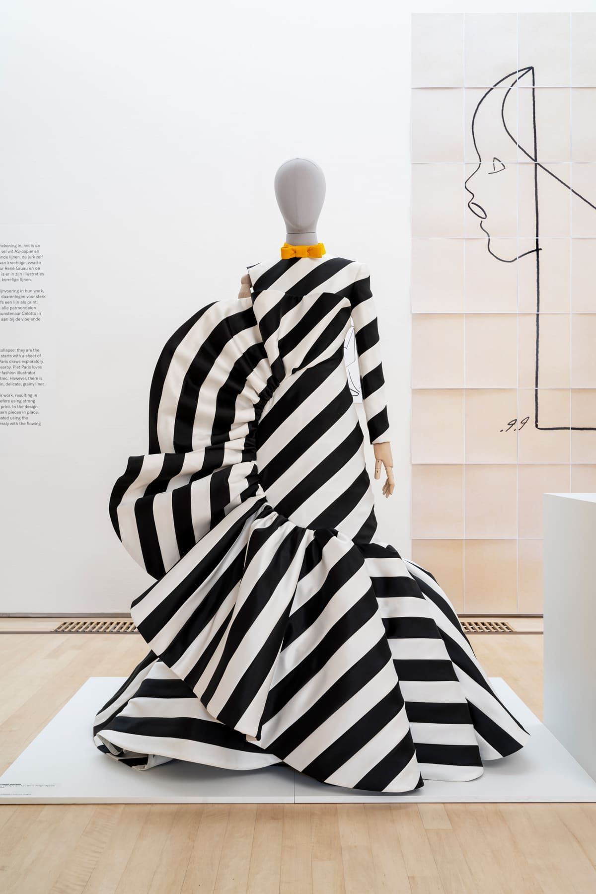 Black and white striped gown on a mannequin, with a ruffled shoulder detail and flared skirt, displayed in a gallery.