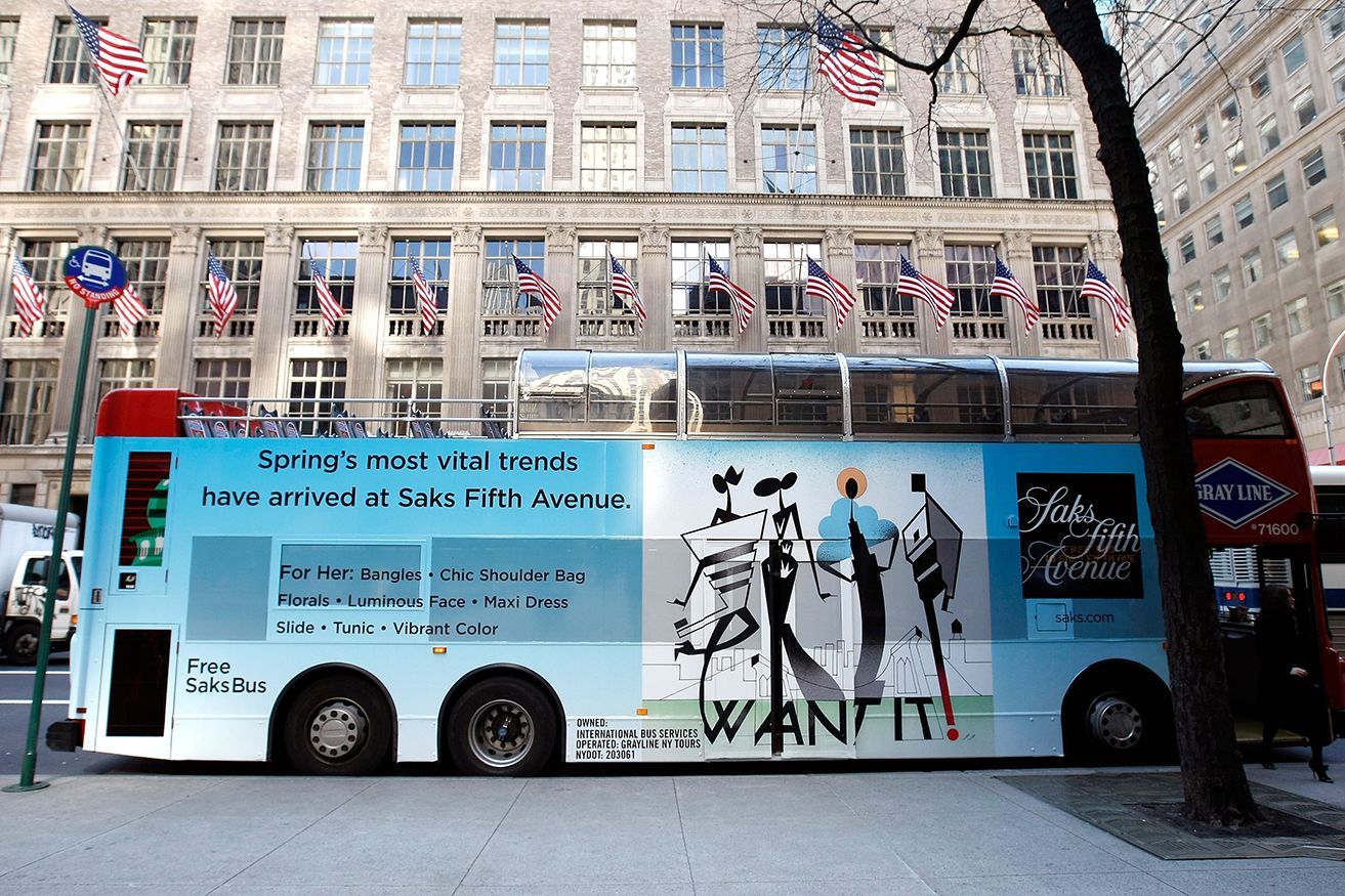 Double-decker tour bus in NYC, with colorful ad graphics, parked on a city street in front of a building with American flags.