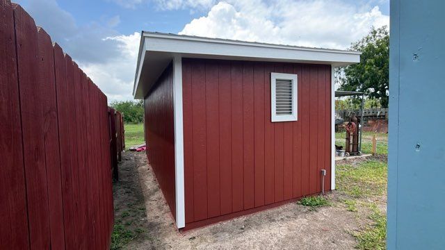Red shed with white trim and a small window, viewed outdoors beside a red fence and blue wall.
