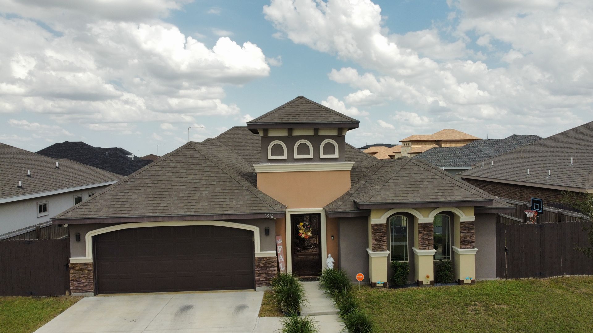 Suburban house with a two-car garage, tan stucco exterior, and dark roof under a cloudy sky