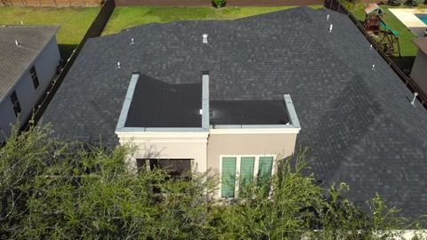 Aerial view of a single-story house with a dark gray roof and beige exterior, surrounded by trees and neighboring homes