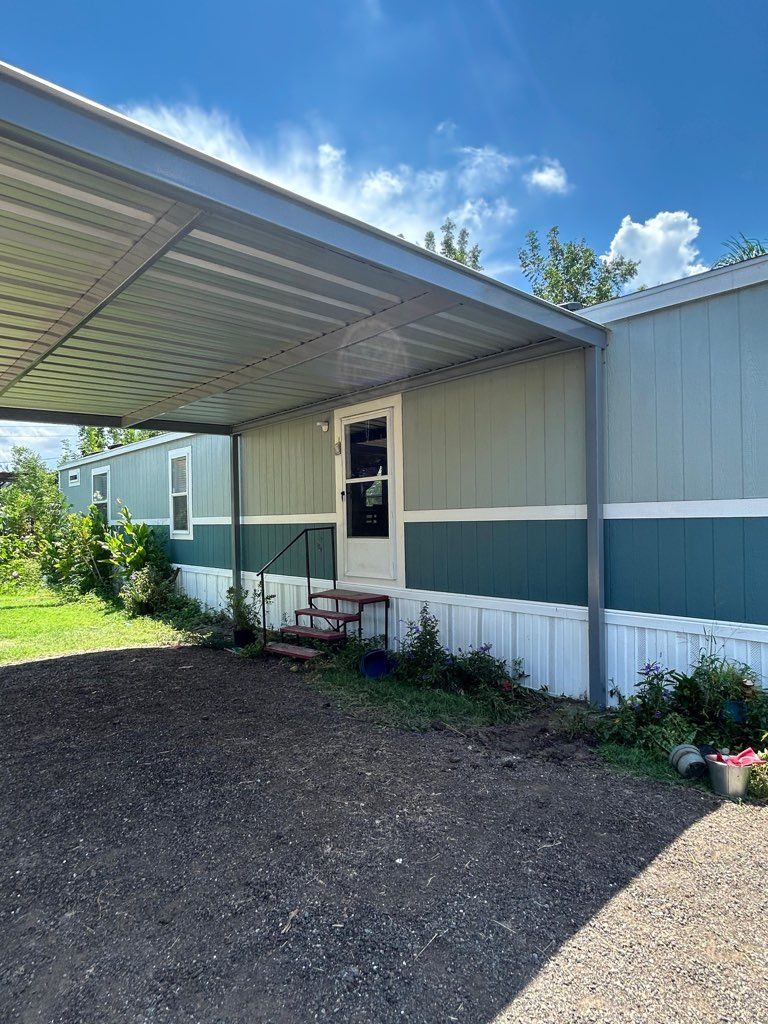 Mobile home exterior with covered carport, white and green siding, and gravel driveway under blue sky