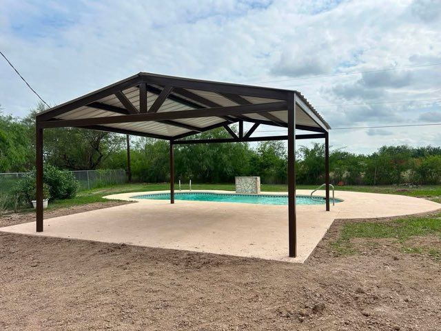 Covered pool under a pavilion in a grassy park area, with a tan deck and cloudy sky.
