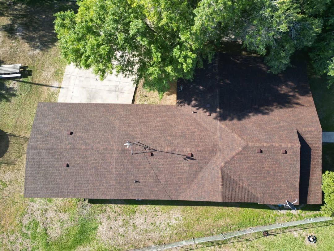 Aerial view of a brown-roofed house with a driveway, lawn, and trees nearby