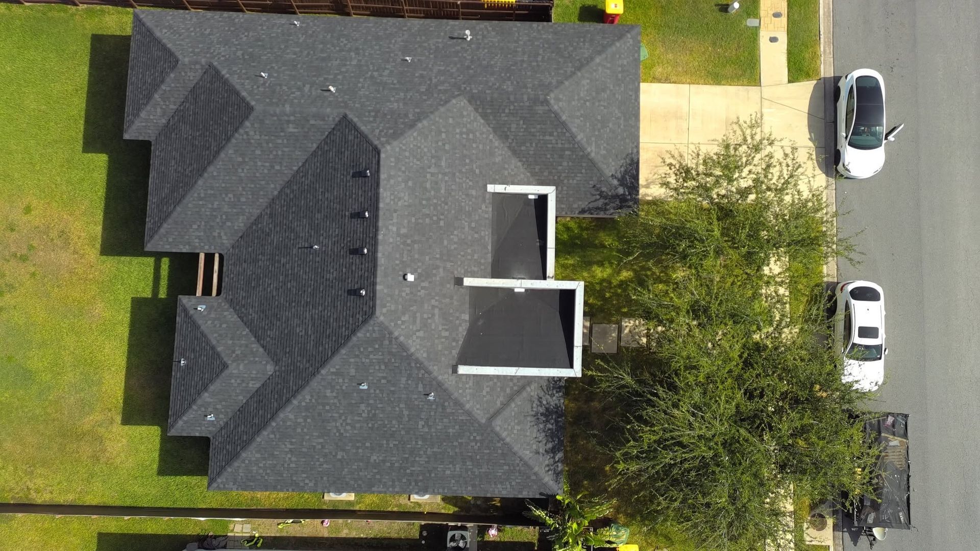 Aerial view of a dark gray house roof with two white dormers and parked cars along the street.